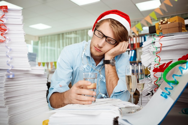 Tired young businessman sitting at workplace among papers on christmas day.
