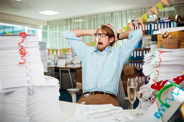 Tired young businessman sitting at workplace among papers on christmas day.