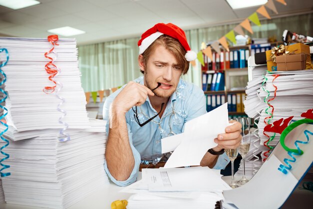 Tired young businessman sitting at workplace among papers on christmas day.