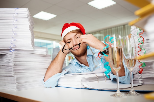 Tired young businessman sitting at workplace among papers on christmas day.