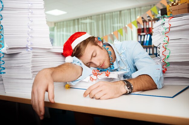 Tired young businessman sleeping at workplace among papers on christmas day.