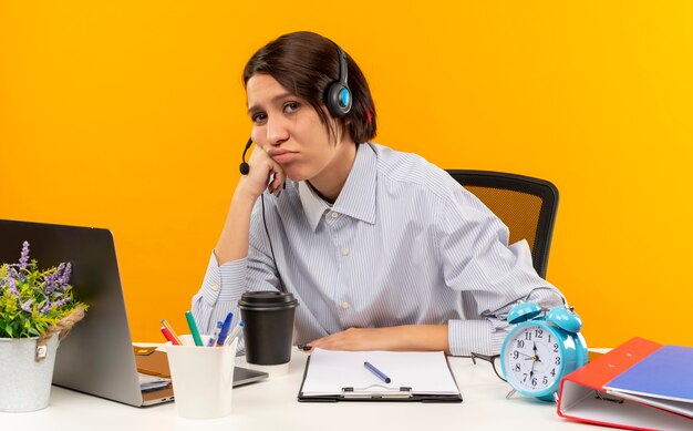 Tired young call center girl wearing headset sitting at desk with work tools putting hand on cheek isolated on orange wall