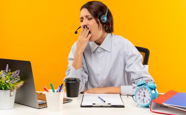 Tired young call center girl wearing headset sitting at desk with work tools yawning with hand on mouth and closed eyes isolated on orange wall