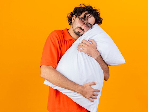 Tired young caucasian ill man wearing glasses hugging pillow putting head on it with closed eyes isolated on orange background with copy space