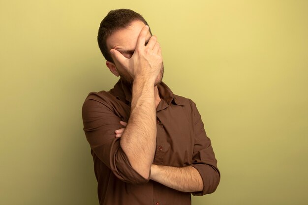 Tired young caucasian man covering face with hand with closed eyes isolated on olive green background with copy space