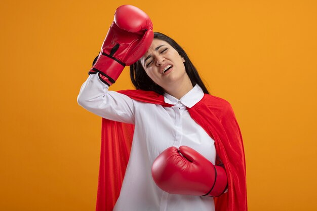 Tired young caucasian superhero girl wearing box gloves touching head with closed eyes isolated on orange background with copy space