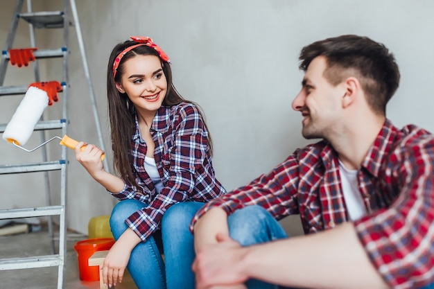 Tired young couple are sitting on the floor with paint brush and bucket while doing repair at home