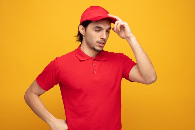 制服とキャップを身に着けている疲れた若い配達人は、黄色の背景で隔離の目を閉じて彼のキャップに触れて腰に手を保ちます