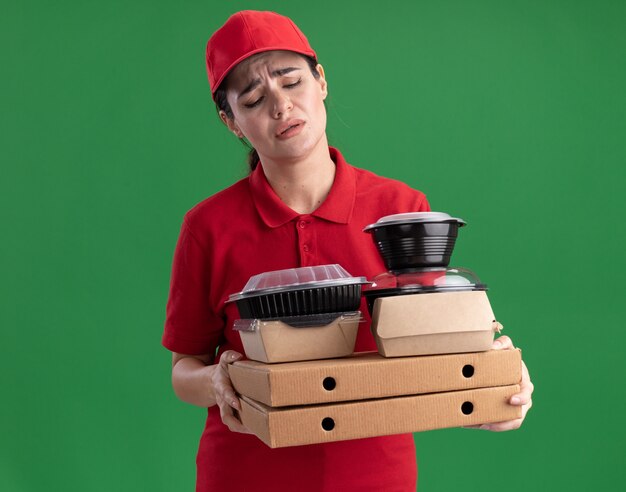 Tired young delivery woman in uniform and cap holding looking at pizza packages with paper food packages and food containers on them isolated on green wall