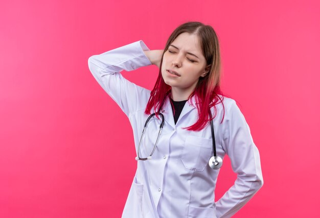 Tired young doctor girl wearing stethoscope medical gown put her hand on neck on pink isolated wall