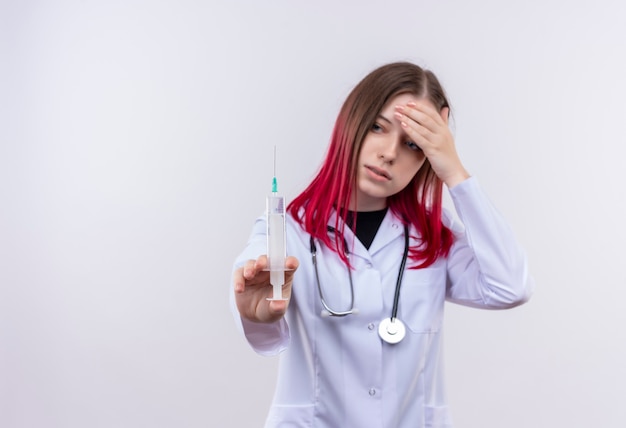 Усталая молодая девушка-врач в медицинском халате со стетоскопом, глядя на шприц на руке, заправляя руку на лоб на изолированной белой стене