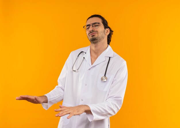 Tired young doctor with medical glasses wearing medical robe with stethoscope holding hands around stomach on isolated yellow wall with copy space