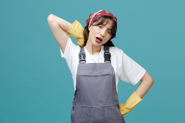 Tired young female cleaner wearing uniform bandana and rubber gloves looking down keeping hand behind neck and on waist isolated on blue background