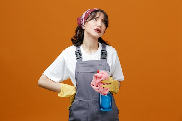 Stanca giovane donna addetta alle pulizie che indossa guanti di gomma uniformi e bandana che tiene uno spolverino di stoffa e un detergente guardando il lato tenendo la mano sulla vita isolata su sfondo arancione