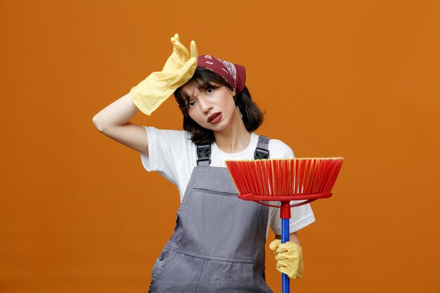 制服のゴム手袋とオレンジ色の背景に分離されたカメラを見て頭に手を保持しているスキージモップを保持しているバンダナを身に着けている疲れた若い女性クリーナー