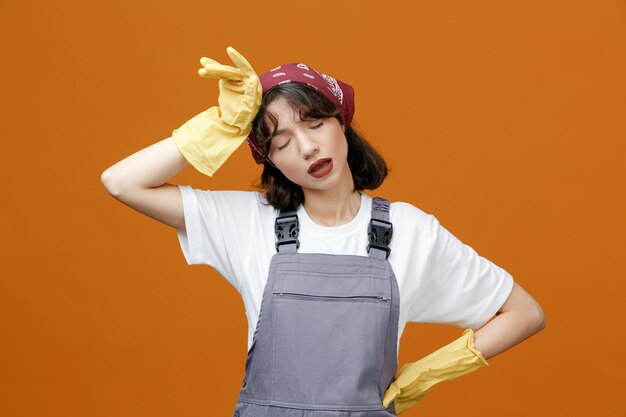 유니폼 고무 장갑과 반다나를 착용하고 머리와 허리에 손을 대고 주황색 배경에 격리된 눈을 감고 지친 젊은 여성 청소부