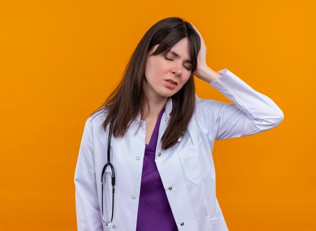 Stanco giovane medico femminile in abito medico con lo stetoscopio tiene la testa con la mano su sfondo arancione isolato con spazio di copia