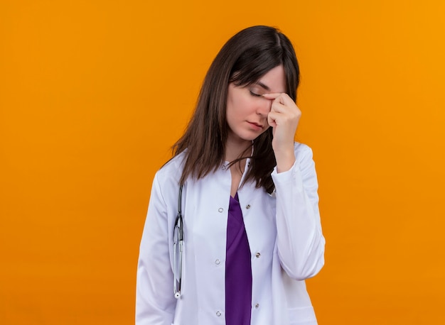 Tired young female doctor in medical robe with stethoscope puts hand on face on isolated orange background with copy space