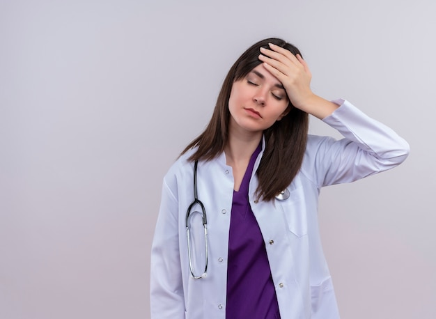 Усталая молодая женщина-врач в медицинском халате со стетоскопом кладет руку на голову на изолированном белом фоне с копией пространства
