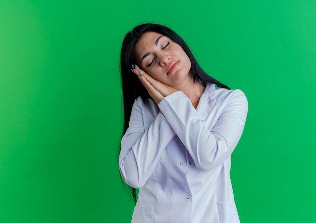 Tired young female doctor wearing medical robe doing sleep gesture with closed eyes 