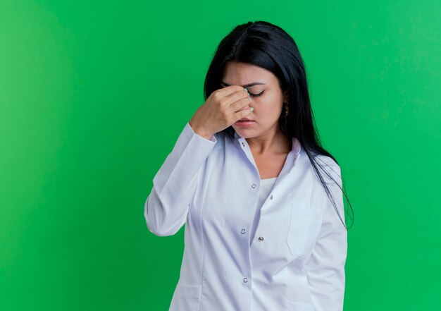 コピースペースのある緑の壁に隔離された目を閉じて鼻を保持している医療ローブを身に着けている疲れた若い女性医師