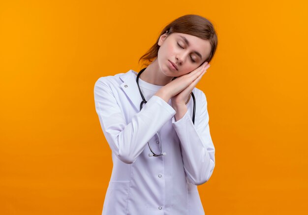 コピースペースのある孤立したオレンジ色の壁で睡眠ジェスチャーをしている医療ローブと聴診器を身に着けている疲れた若い女性医師