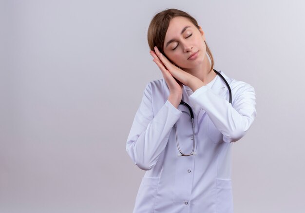 Tired young female doctor wearing medical robe and stethoscope doing sleep gesture on isolated white wall with copy space