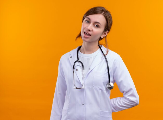 Tired young female doctor wearing medical robe and stethoscope putting hand behind her back on isolated orange wall with copy space