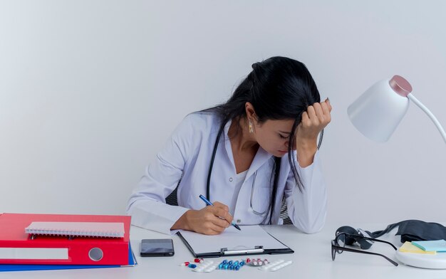 Усталая молодая женщина-врач в медицинском халате и стетоскопе сидит за столом с медицинскими инструментами, держа ручку, положив руку на голову с закрытыми изолированными глазами