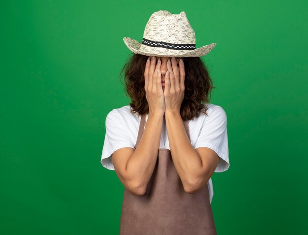 Tired young female gardener in uniform wearing gardening hat covered face with hands