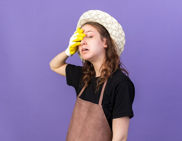 Tired young female gardener wearing gardening hat with gloves covered eye with hand 