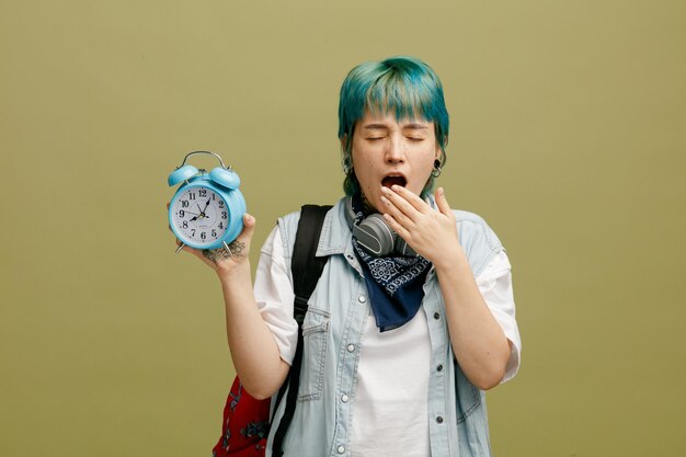 Free photo tired young female student wearing headphones and bandana on neck and backpack showing alarm clock keeping hand in front of mouth yawning with closed eyes isolated on olive green background