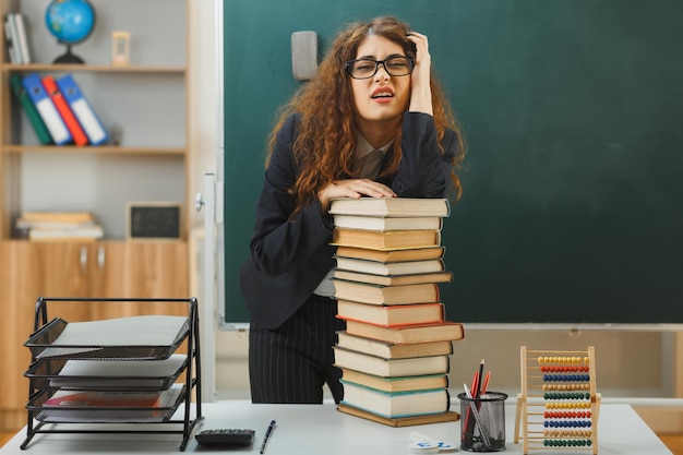 tired young female teacher standing in front blackboard holding books in classroom