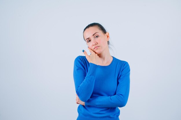 Tired young girl is looking at camera by holding hand on cheek on white background