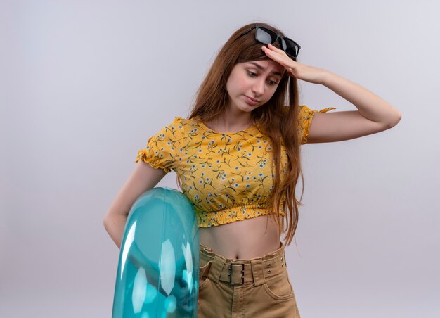 Tired young girl wearing sunglasses on head holding swim ring putting hand on head looking down on isolated white wall