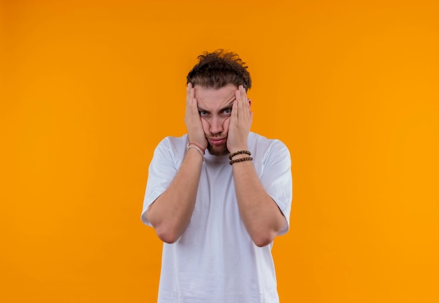 Tired young guy wearing white t-shirt put his hands on cheeks on isolated orange wall