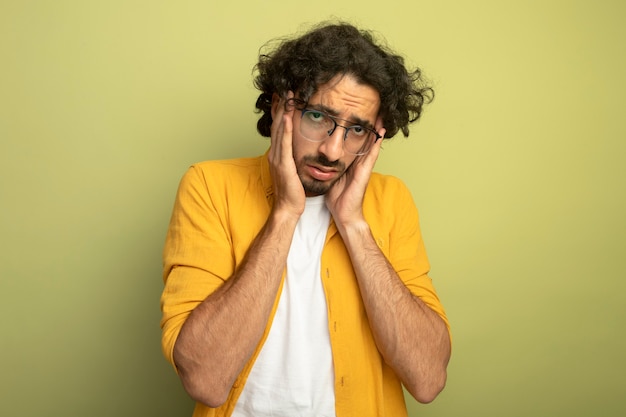 Tired young handsome caucasian man wearing glasses putting hands on head looking at camera isolated on olive green background with copy space