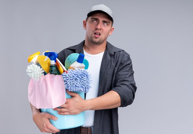 Tired young handsome cleaning guy wearing t-shirt and cap holding bucket of cleaning tools isolated on white wall with copy space