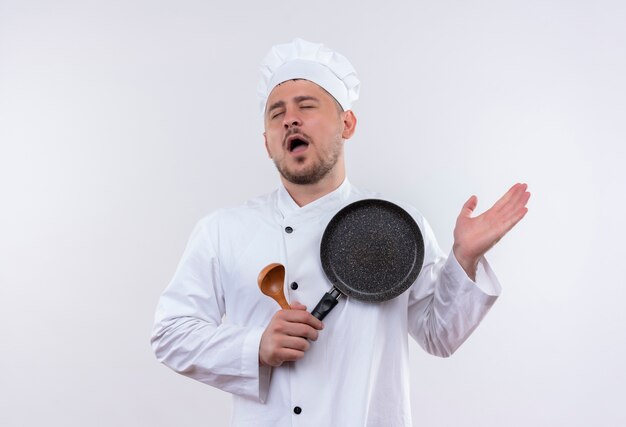 Усталый молодой красивый повар в униформе шеф-повара, держащий ложку и сковороду, показывает пустую руку с закрытыми глазами, изолированными на белом пространстве