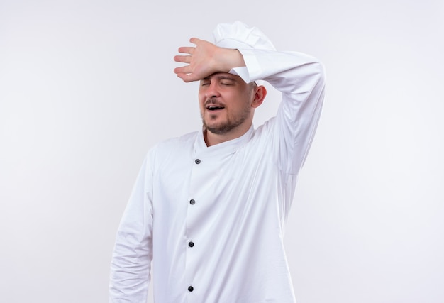 Tired young handsome cook in chef uniform putting hand on forehead and yawning with closed eyes isolated on white space