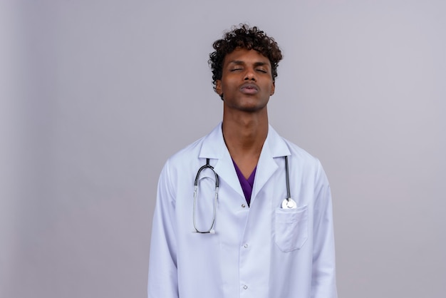 A tired young handsome dark-skinned male doctor with curly hair wearing white coat with stethoscope closing his eyes and thinking 