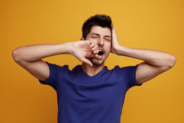 Tired young handsome man looking at camera keeping hands on head and near mouth yawning on yellow background