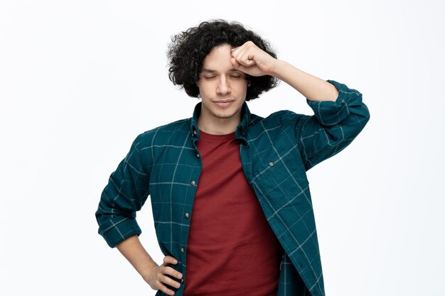 Tired young handsome man touching head with fist while keeping hand on waist with closed eyes isolated on white background