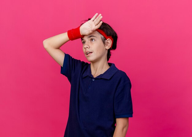 Tired young handsome sporty boy wearing headband and wristbands with dental braces keeping hand on forehead looking up isolated on crimson background with copy space