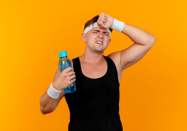 Tired young handsome sporty man wearing headband and wristbands holding water bottle and putting hand on forehead isolated on orange background