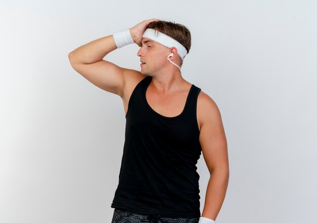 Tired young handsome sporty man wearing headband and wristbands with headphones putting hand on head with closed eyes isolated on white background with copy space