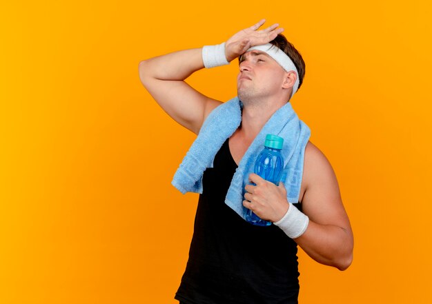 Tired young handsome sporty man wearing headband and wristbands with towel around neck looking up with hand on forehead holding water bottle isolated on orange background with copy space