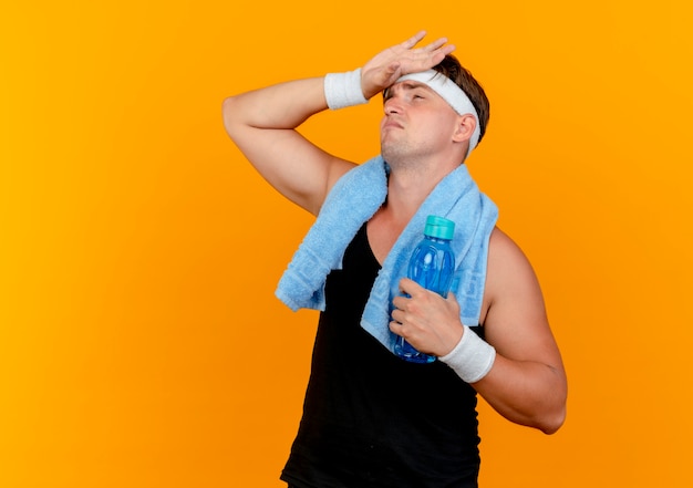 Усталый молодой красивый спортивный мужчина с повязкой на голову и браслетами с полотенцем на шее, глядя вверх рукой на лбу, держа бутылку с водой, изолированную на оранжевом фоне с копией пространства