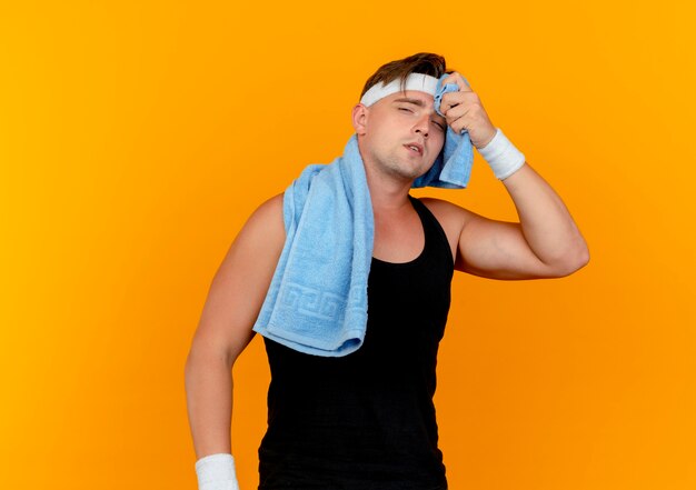 Усталый молодой красивый спортивный мужчина с повязкой на голову и браслетами с полотенцем на шее, вытирая пот полотенцем, изолированным на оранжевом фоне