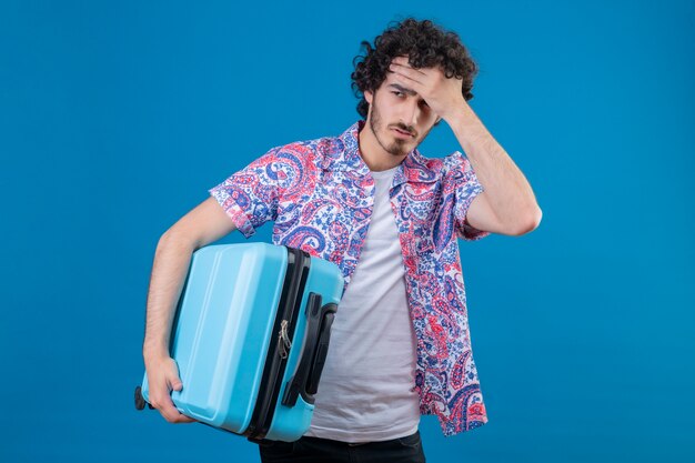 Усталый молодой красивый путешественник мужчина держит чемодан рукой на лбу на изолированной синей стене с копией пространства